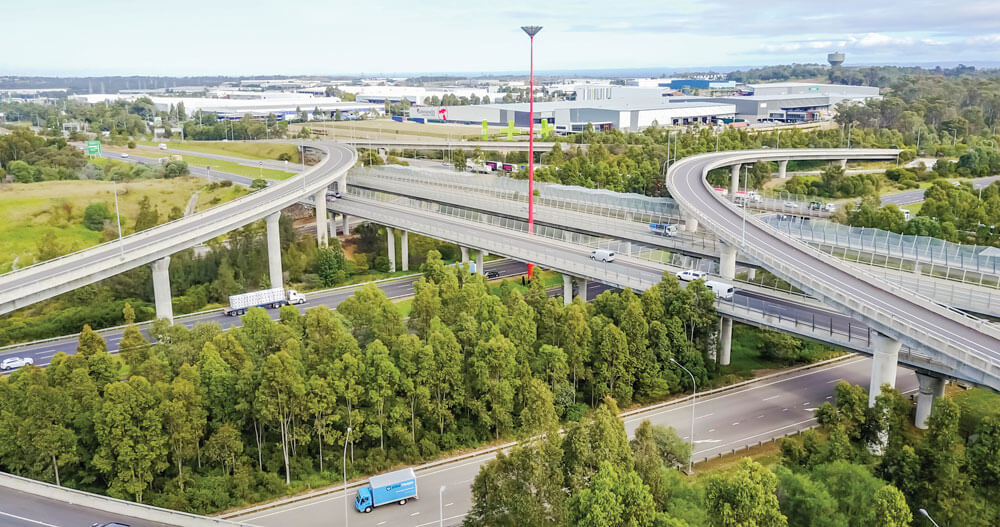View of motorway intersection interspersed with sections of trees with buildings and sky in the distance
