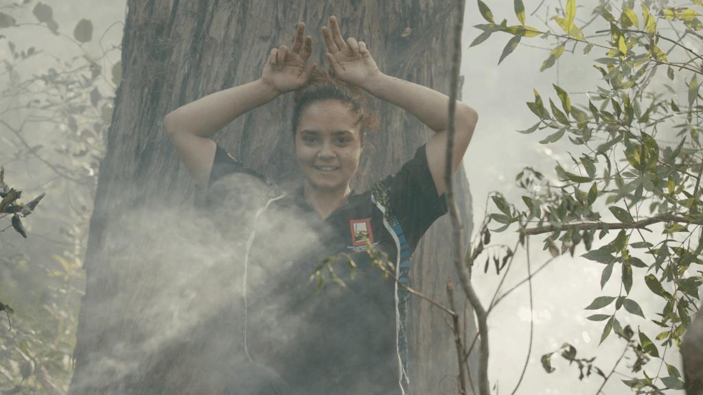 Aboriginal girl holds hands above head surrounded by smoke